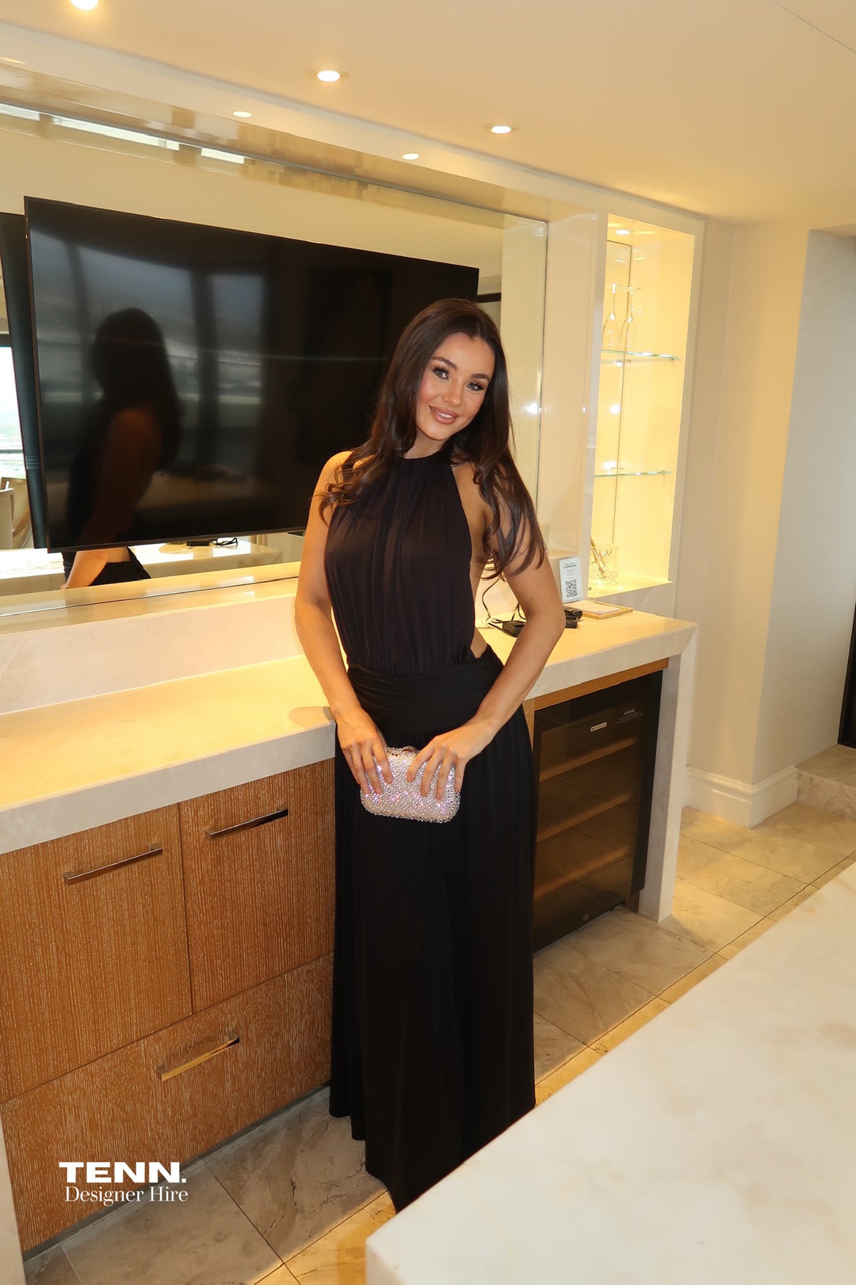 Arcina Ori Santina Black Dress available for hire at TENN Designer Hire — effortlessly elegant gown with halter neckline, flowing bodice, waist band, open back, and flowing skirt, perfect for formal occasions and styled with gold accessories.