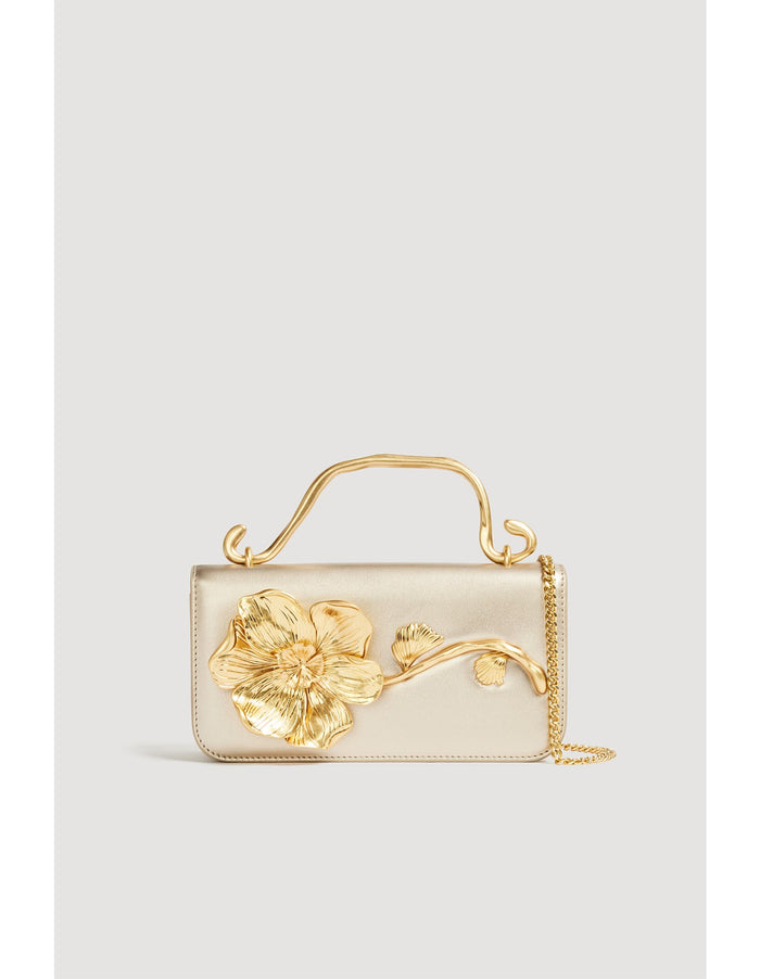 Aje Celeste Flower Clutch featuring a gold top handle and gold sculpted flower detail, photographed against a clean studio backdrop.